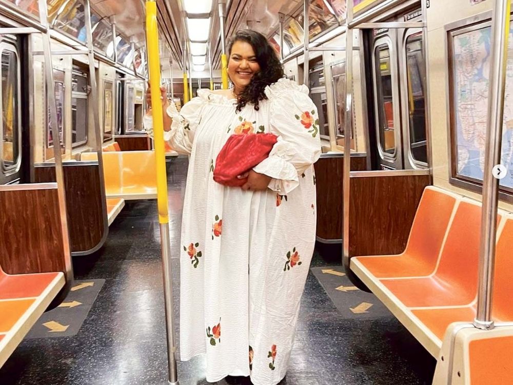 A smiling person in a white floral dress holds a red clutch while standing in an empty subway car.