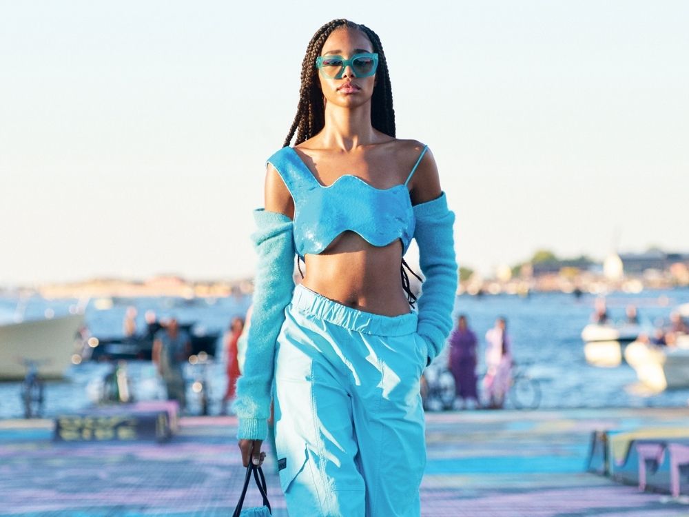A model walks on a seaside runway wearing a bright blue outfit with a cropped top and pants, exuding confidence. The scene is set against a sunny, coastal backdrop.