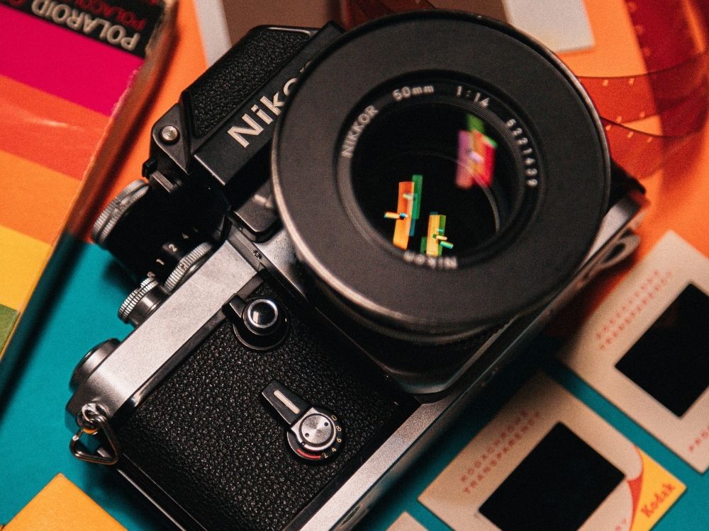 A vintage Nikon camera with a 50mm lens is surrounded by colorful items, including a Polaroid box and film slides, creating a nostalgic, artistic vibe.