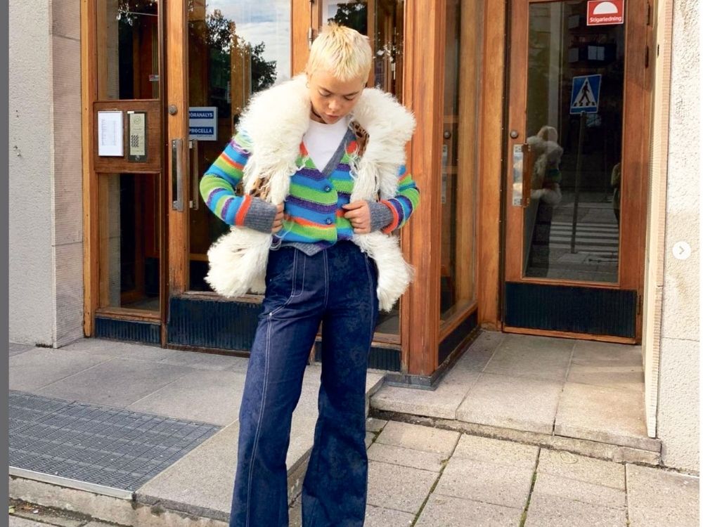 Person stands outside a wooden door, wearing a colorful striped sweater, fluffy vest, and dark jeans, looking down in a casual and stylish pose.