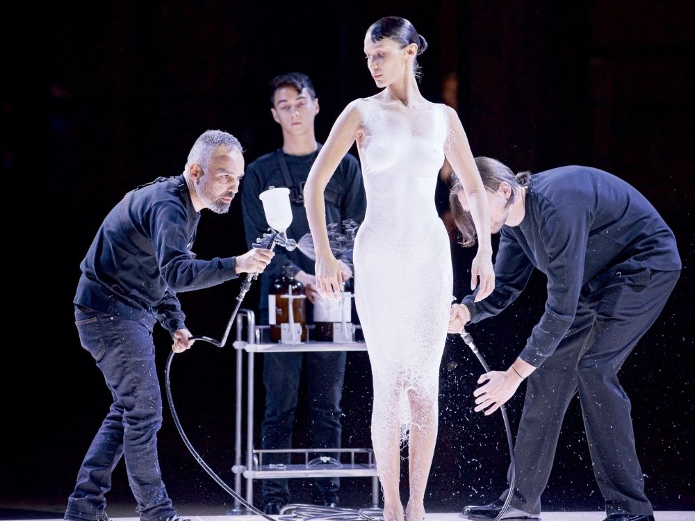 Model standing while two designers spray a white dress onto her using an airbrush. The scene is artistic and innovative.