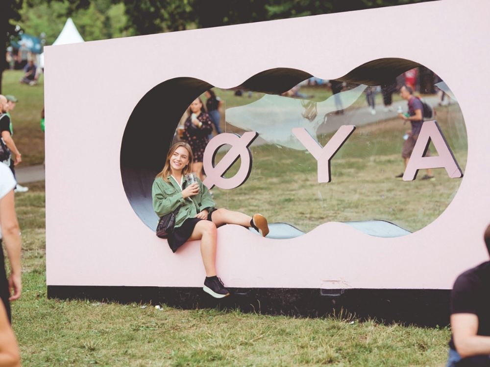 A woman sits playfully inside a large pink sign with the letters "ØYA" at a park festival. She smiles, holding a drink.