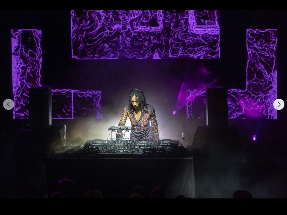 A DJ performs energetically behind a mixing console against a backdrop of vivid purple abstract patterns. The scene is lively and dynamic.