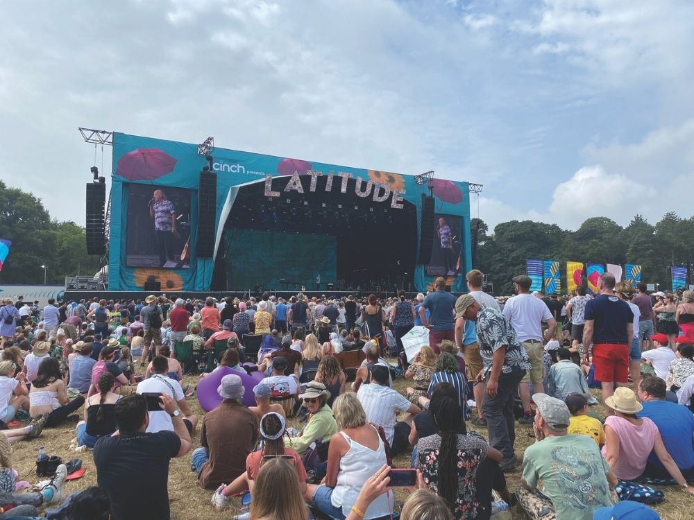 Crowd gathered at an outdoor music festival under a partly cloudy sky, facing a large stage with "LATITUDE" sign.