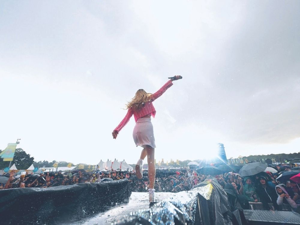 A performer in a pink outfit faces a cheering crowd while holding a microphone, under a dramatic sky. Excited fans hold umbrellas in a festival setting.