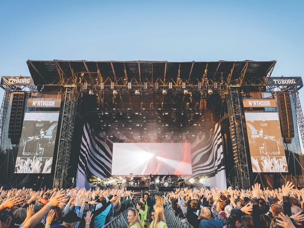Outdoor music festival at dusk with a large stage and vibrant lights, featuring "Northside" branding. A packed, cheering crowd raises hands.