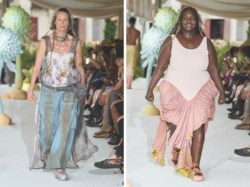 Two models walk on a fashion runway. The left model wears a floral top and blue skirt, the right model wears a pink dress. Both look confident and joyful.