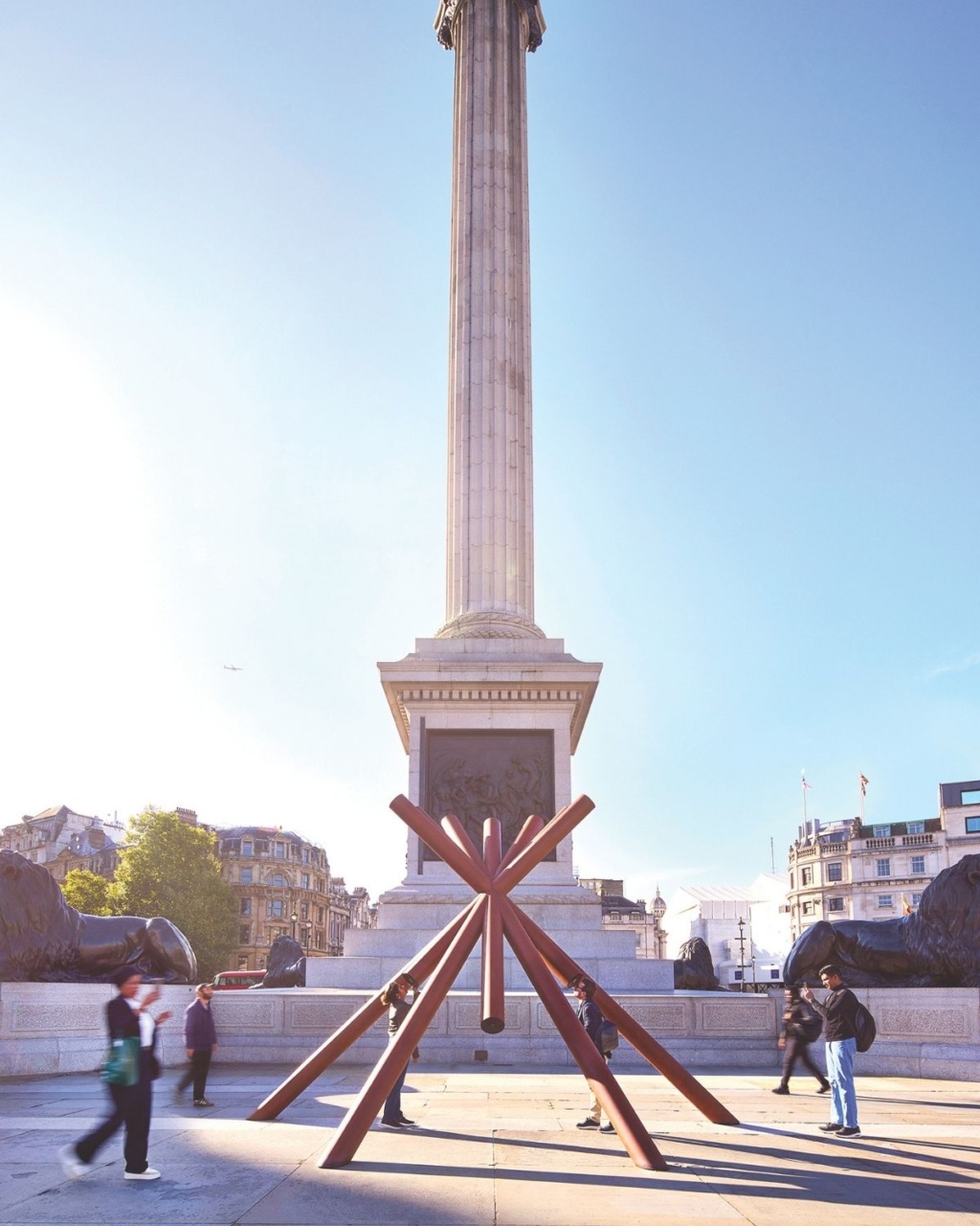 A reddish-brown, multi-pronged modern art sculpture positioned in the foreground and people walking around the plaza in bright daylight.