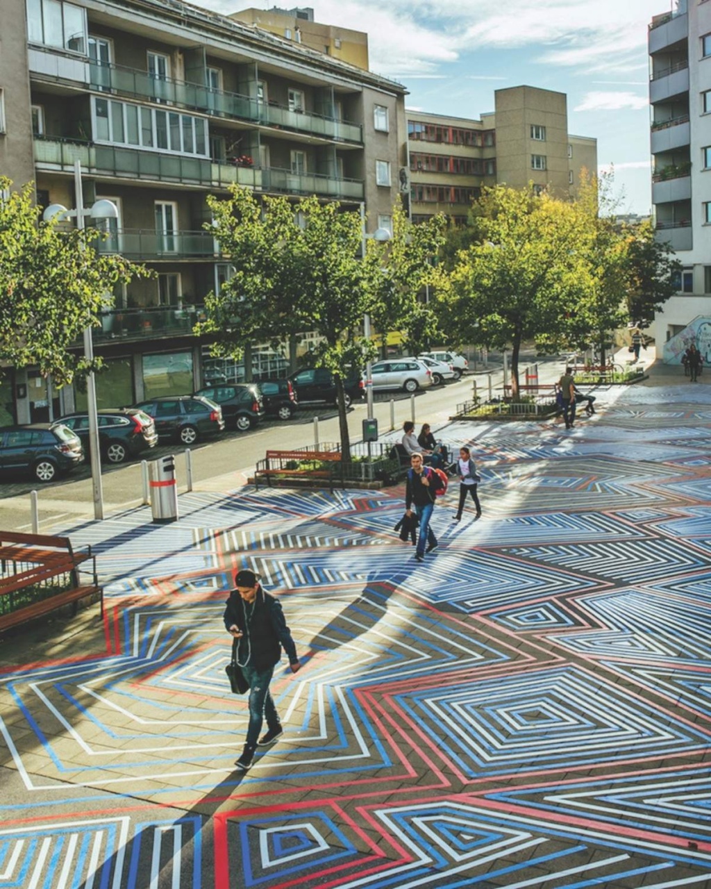 A brightly colored square with intricate geometric street art patterns, surrounded by apartment buildings, trees, and parked cars under a partly cloudy sky.