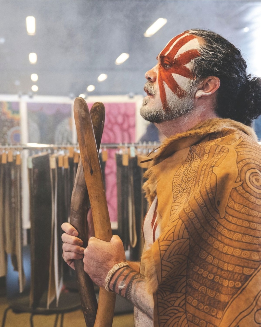 An Indigenous man with red facial paint and patterned leather garments stands holding a pair of wooden clapsticks inside a gallery displaying colorful artworks.