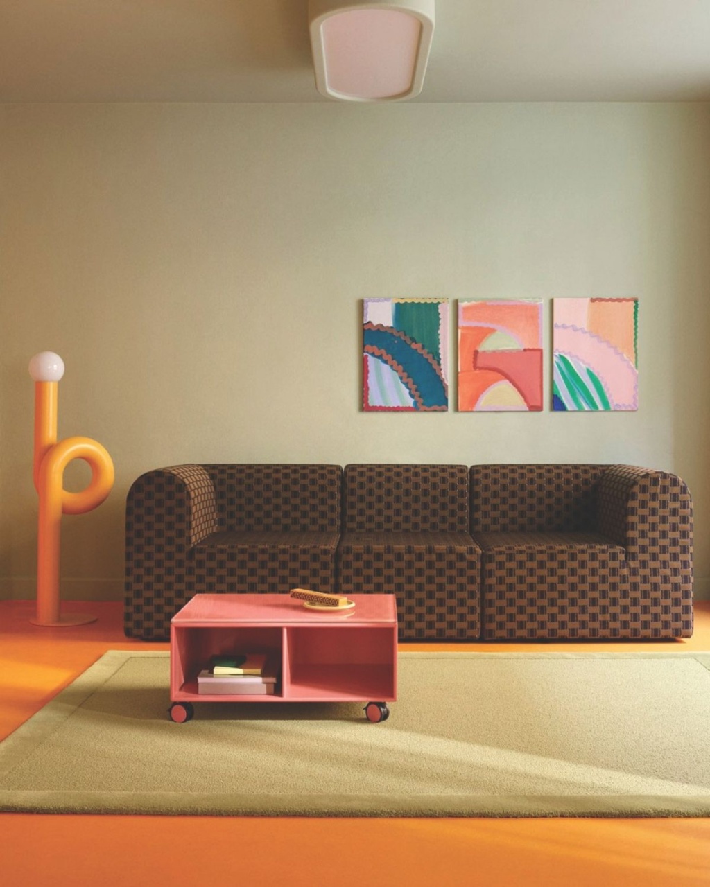 A low-profile sofa with a dark brown checkered pattern, set against a warm beige wall with three abstract paintings, and a bright orange floor with a pale green rug, while a pink rolling coffee table sits in the foreground.