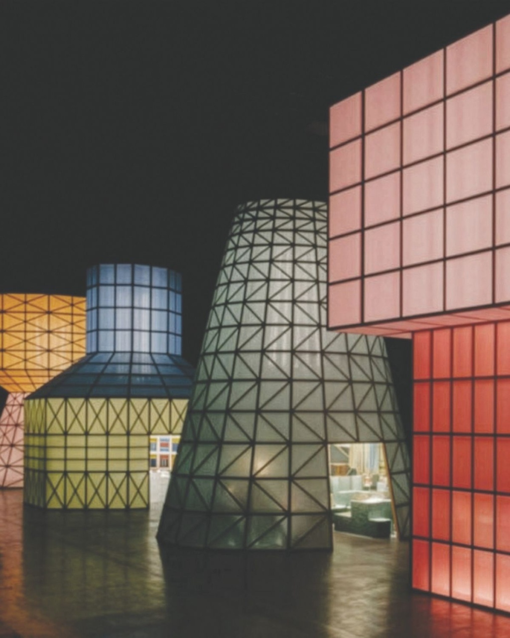 Futuristic art installation featuring several large, illuminated geometric structures in various shapes and pastel colors against a dark background.