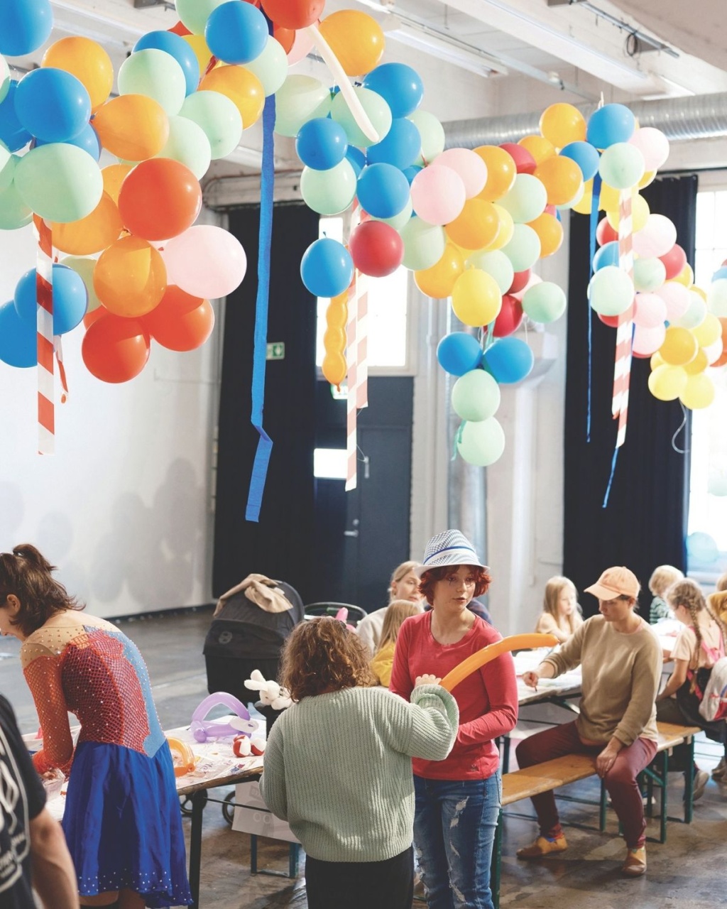 Children and adults gather around tables in a large room decorated with colorful balloons overhead, participating in crafts and activities.