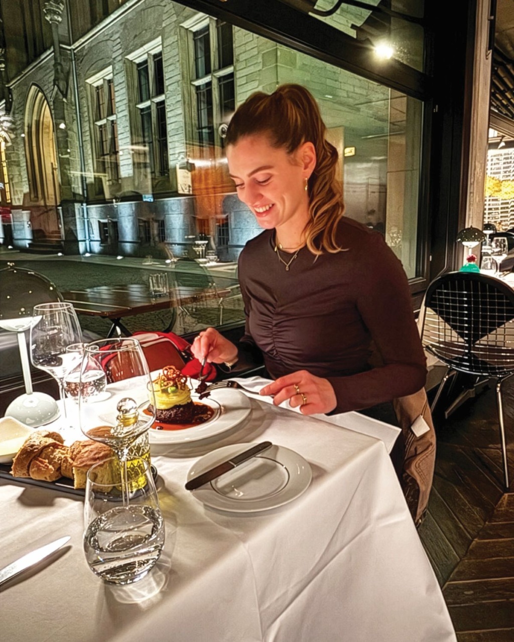 Smiling woman enjoys dessert at a white-tablecloth restaurant, surrounded by wine glasses and pastries, with a historic building visible outside.