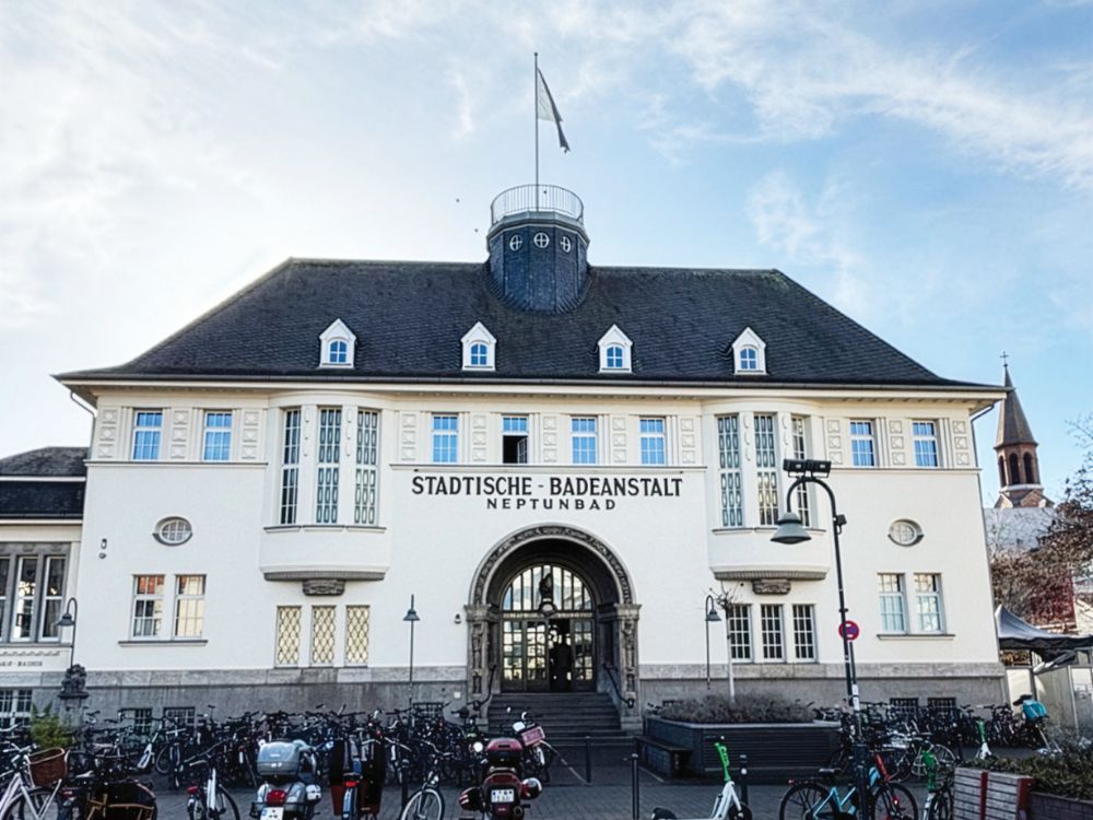 Historic white German bathhouse marked “Städtische Badeanstalt,” with ornate windows, tower, flag, bicycles, and a calm city-street scene.
