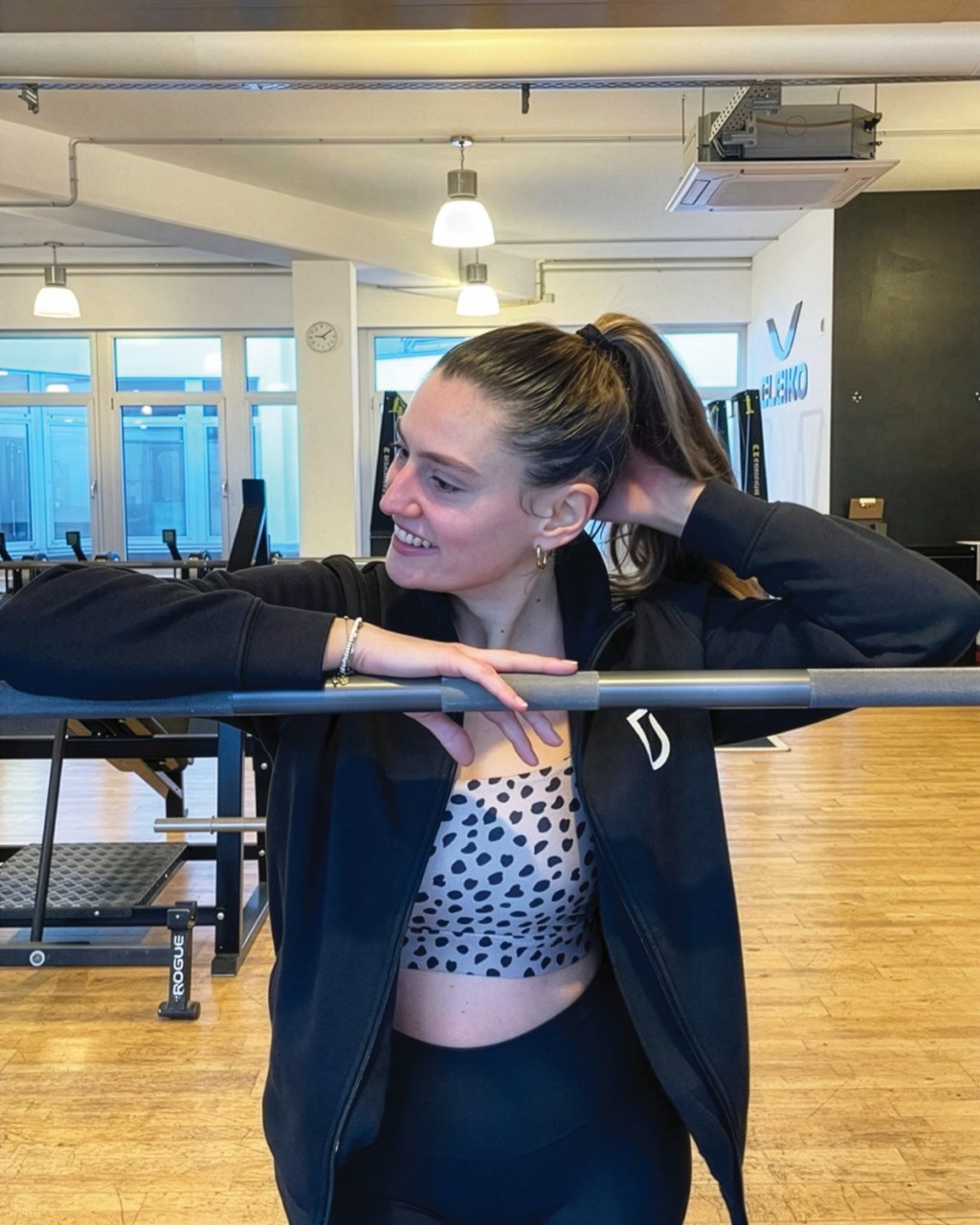 Smiling woman in workout clothes leans on a barre in a bright sports studio, adjusting her ponytail.