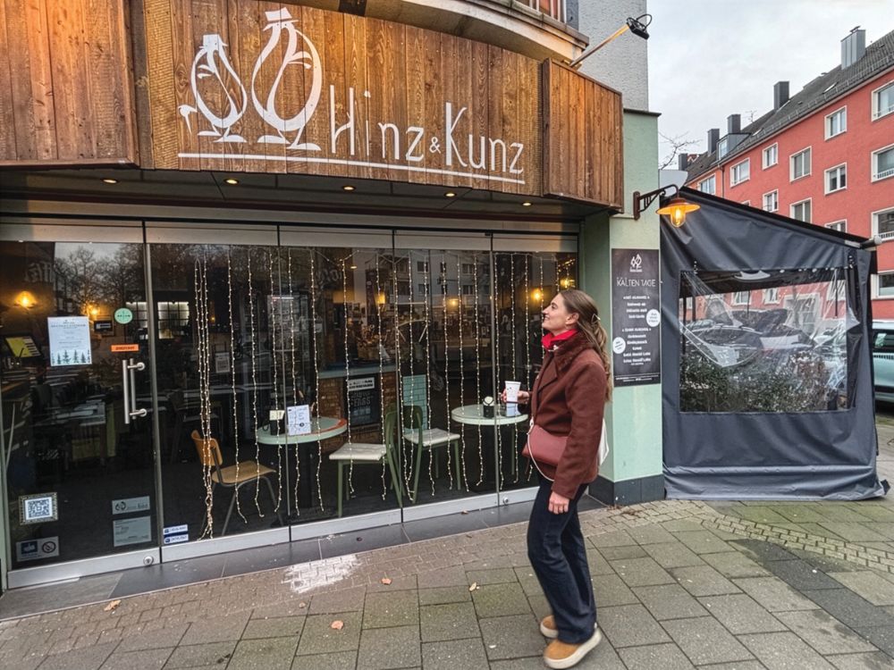 Person in a red scarf stands outside the Hinz & Kunz café, with warm lights, wood signage, and a covered patio on a city street.