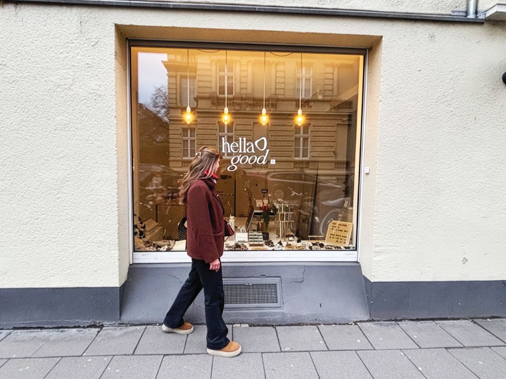 Woman walks past a warmly lit shop window displaying “hella good,” with clothing and décor; building reflections create a cozy urban scene.