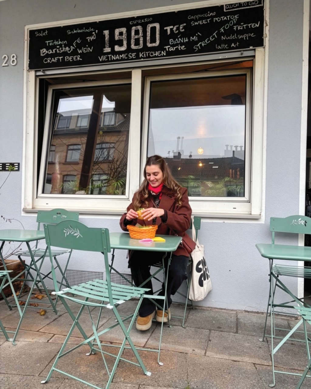 Woman enjoying food at an outdoor café, seated at a green table beneath a “1980 Vietnamese Kitchen” sign; cozy urban street scene.