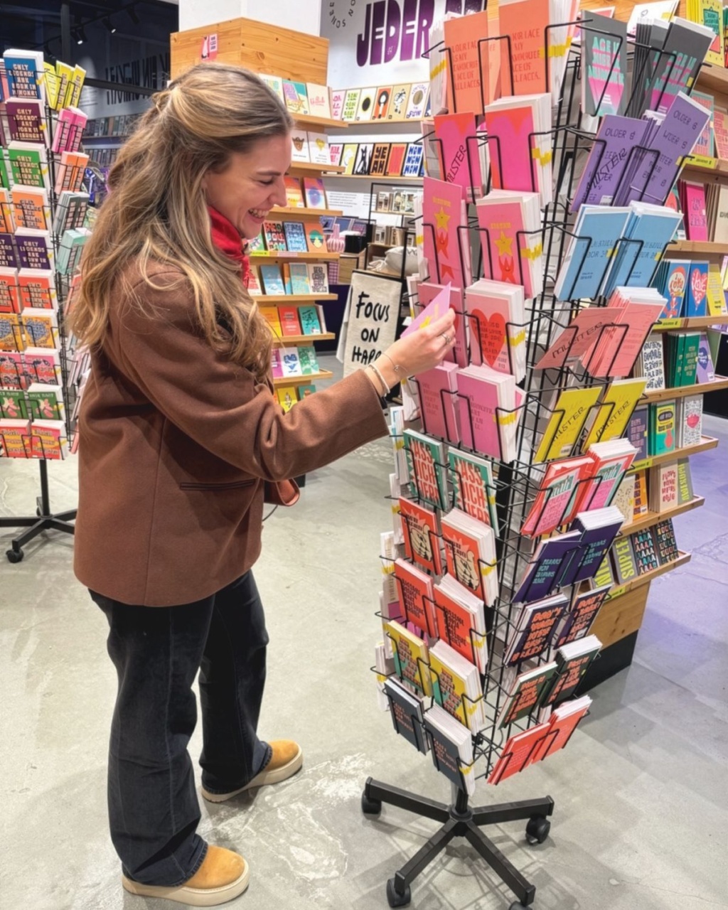Woman browses colorful greeting cards on a rotating display in a bookstore, smiling as she examines a pink card among many designs.