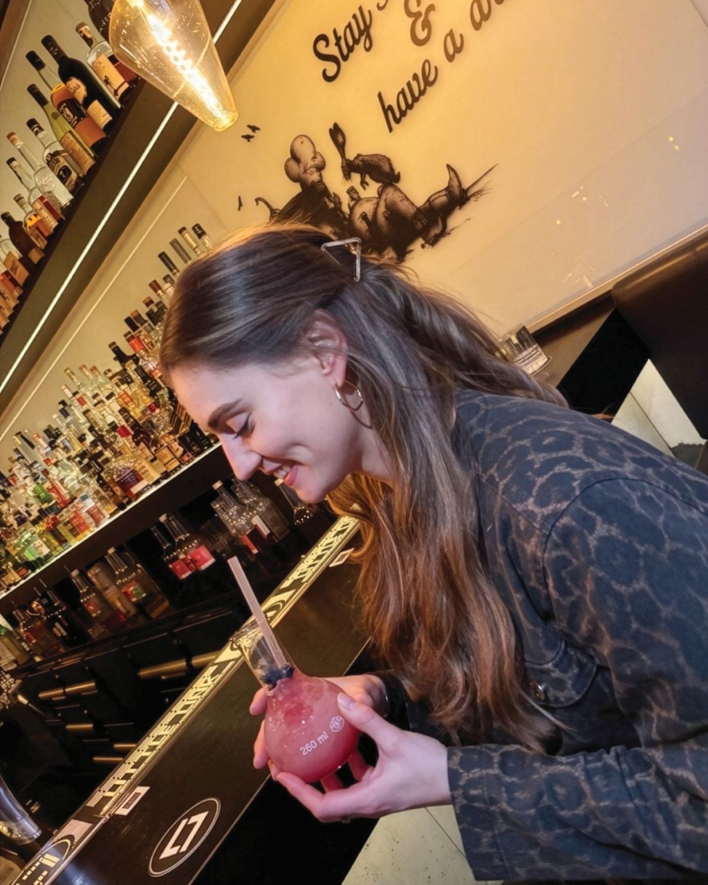 Smiling woman in a leopard-print jacket holds a pink cocktail at a warmly lit bar, surrounded by liquor bottles and whimsical wall art.