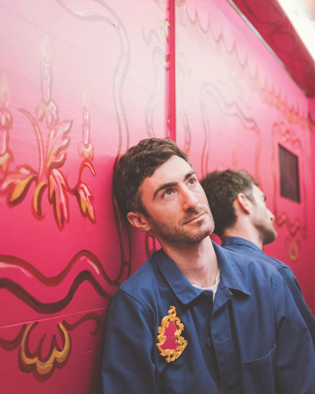 A thoughtful man in a blue uniform with a gold crest leans against an ornate pink wall, gazing upward as his reflection appears in its glossy panels.