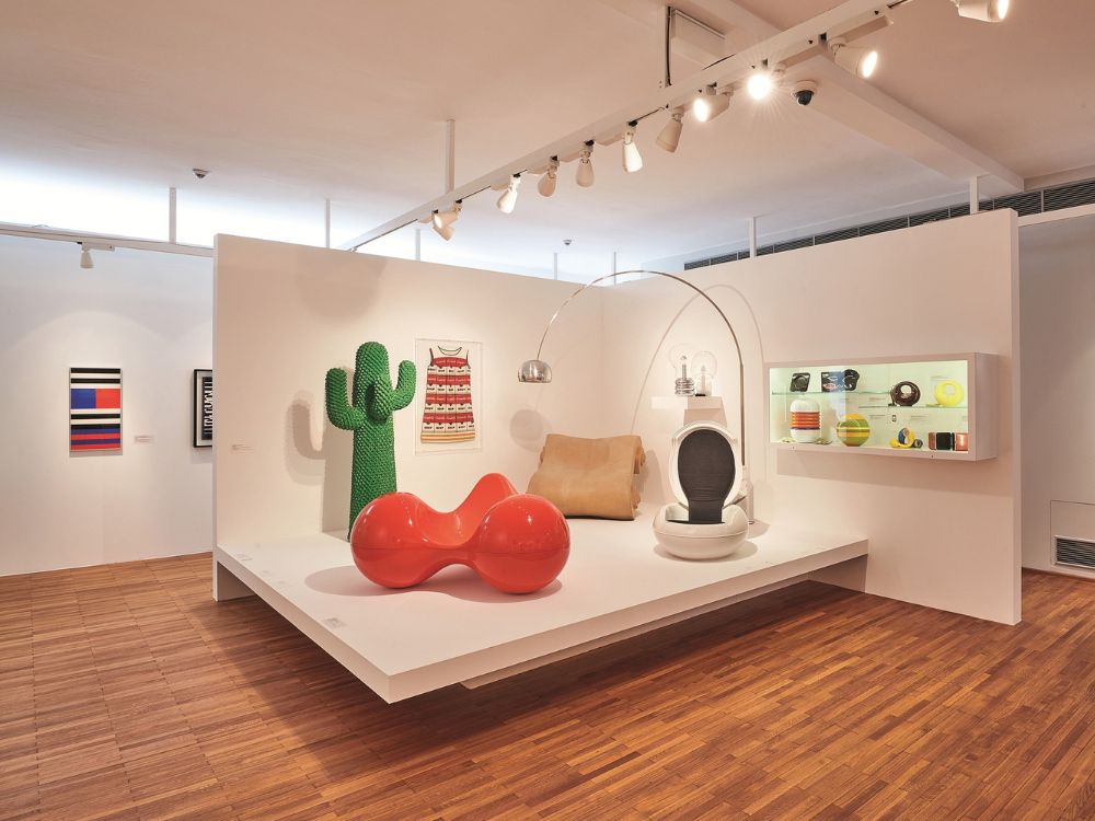 A bright design gallery displays a red sculptural sofa, cactus-shaped chair, suspended egg seat, and colorful objects on a white platform.