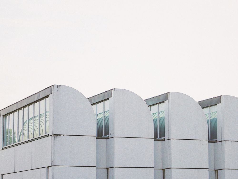 A row of minimalist white modern buildings with rounded upper facades and tall glass windows stands beneath an overcast sky.