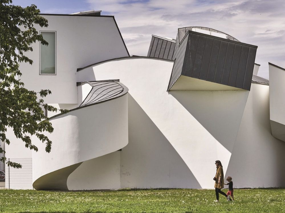 Sculptural white modernist building with bold angular forms and dark rooftop windows; a woman and child walk across the lawn.