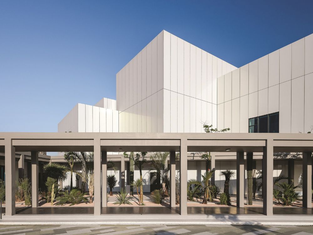 Modern white geometric building with a shaded colonnade, landscaped courtyard, and reflective pool under a clear blue sky; calm, minimalist architecture.