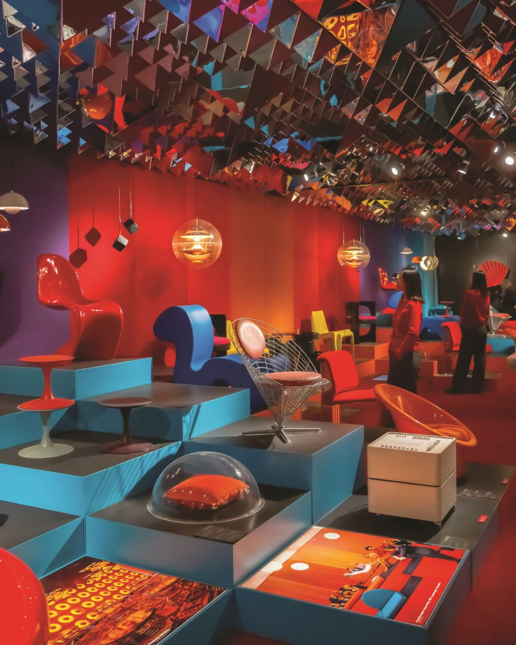 Vibrant retro-futuristic design showroom with sculptural chairs, glowing pendant lights, mirrored ceiling panels, and visitors exploring colorful displays.