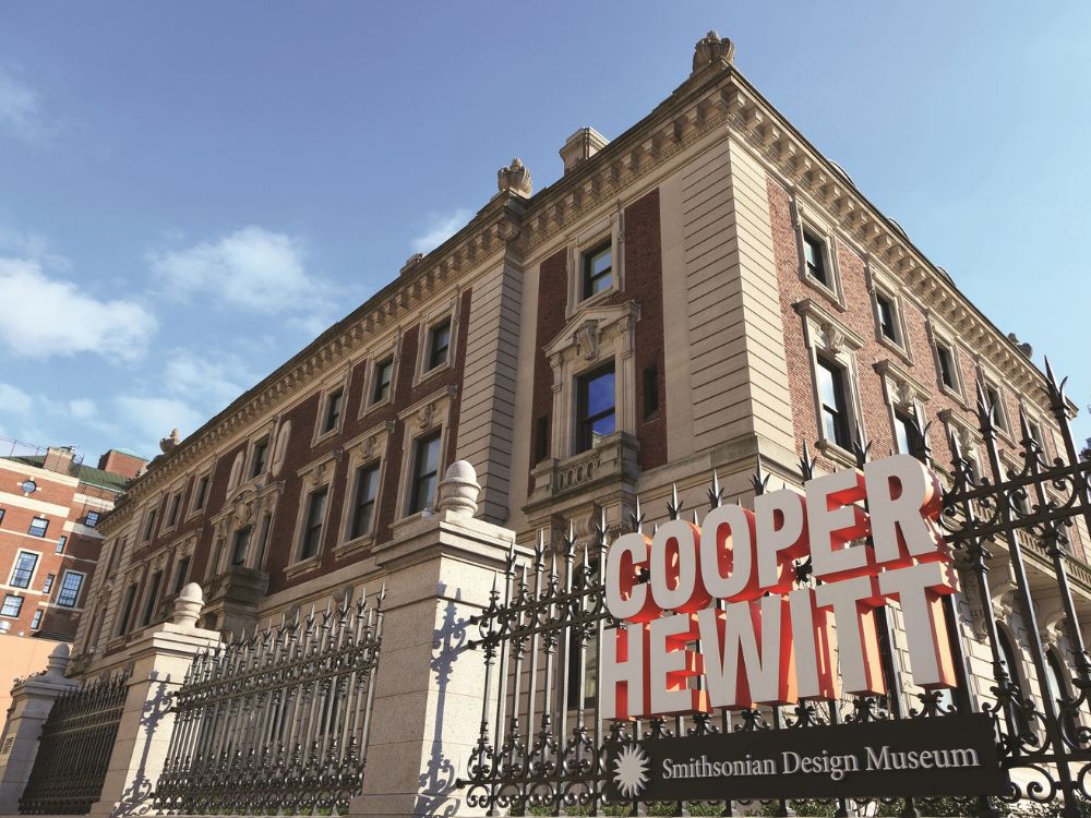 Ornate Cooper Hewitt Smithsonian Design Museum building behind an elaborate iron fence, its bold red sign prominent in bright sunlight.