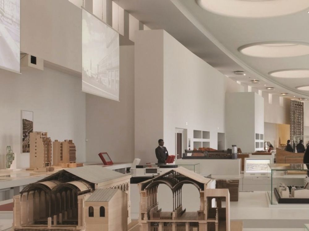 An airy museum gallery displays architectural models on broad tables, with visitors nearby beneath high curved ceilings and soft neutral lighting.