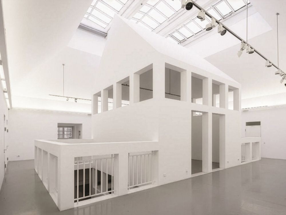 Sunlit, minimalist white gallery with a sculptural two-level structure, tall rectangular openings, and skylights.
