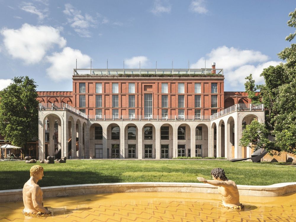 A red-brick building with tall white arches overlooks a lawn and shallow pool, where two sculptural figures face each other in a calm.
