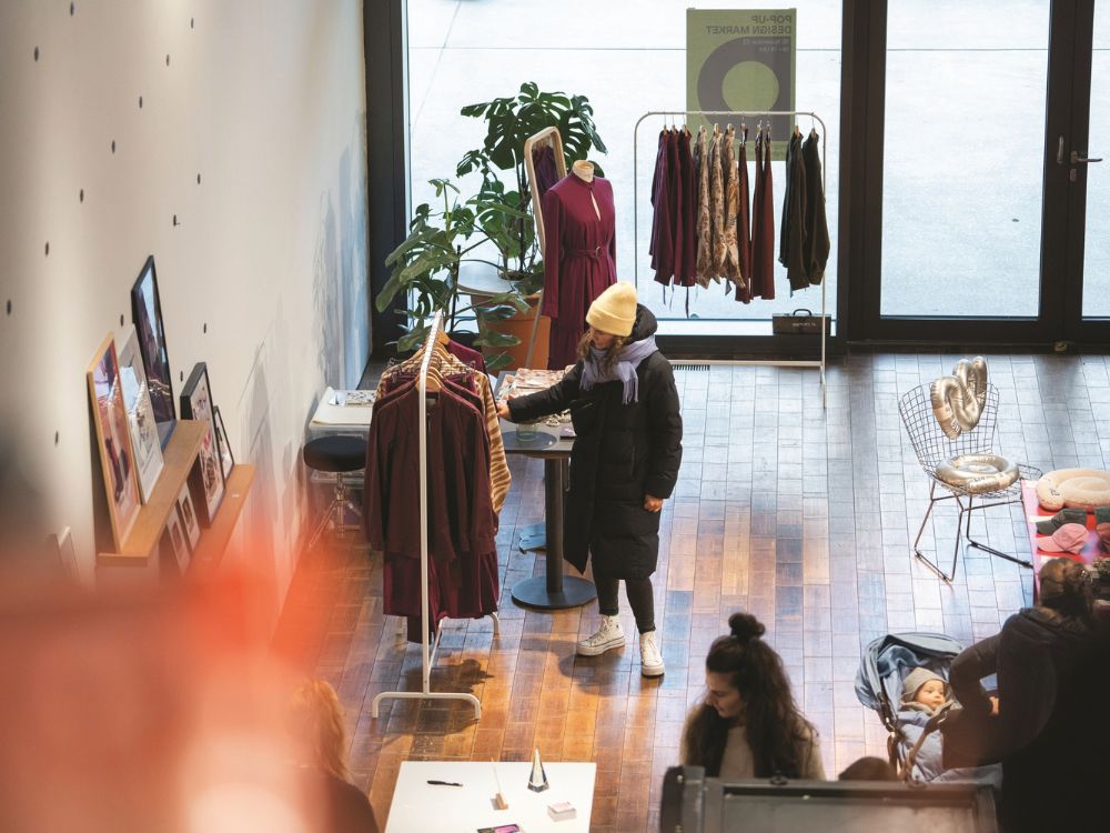 In a sunlit boutique, a shopper in a yellow beanie browses a clothing rack while another person sits nearby.