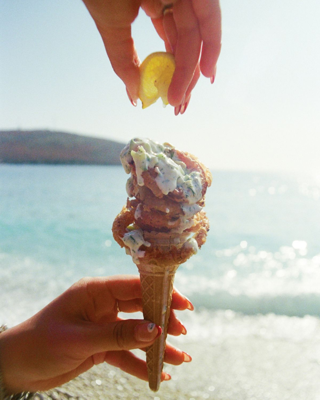 A hand holds a tall, melting ice cream cone by the sunny beach as another hand squeezes lemon over it.