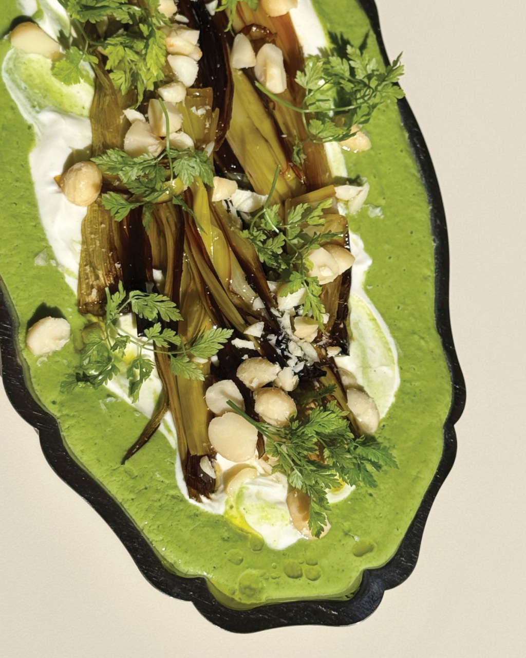 Overhead view of charred leeks on bright green herb sauce, topped with fresh herbs and chopped nuts on a black platter.