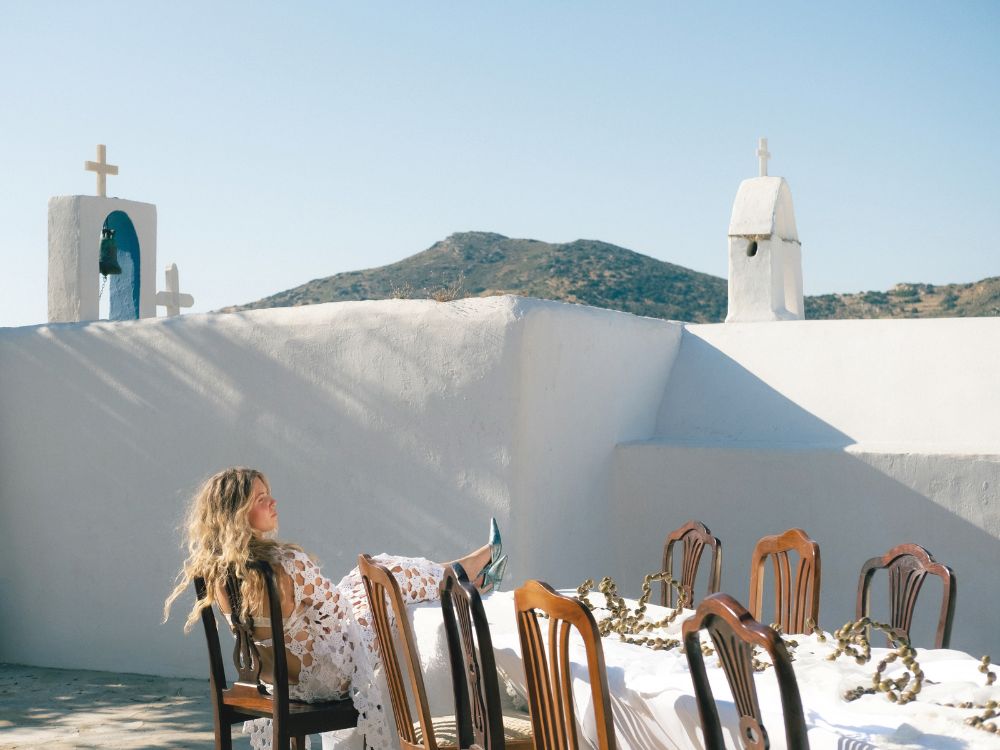 A woman in a white lace dress sits among wooden chairs on a sunlit rooftop terrace, with white chapels, crosses, and a mountain behind her.