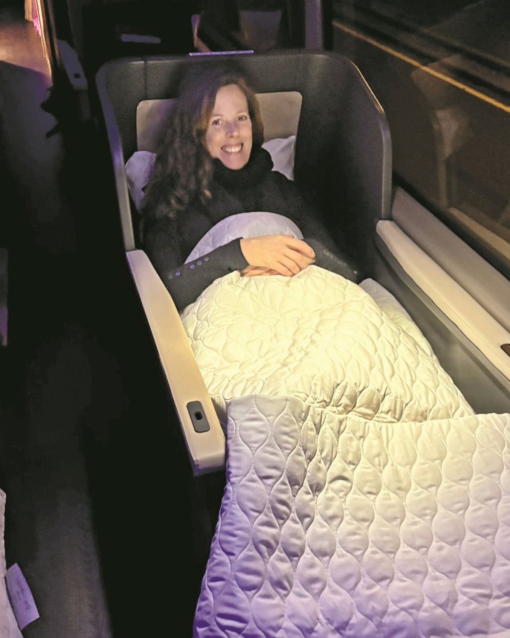 A smiling woman reclines in a cushioned sleeper seat on a train, tucked beneath a quilted blanket, creating a cozy, relaxed nighttime travel scene.