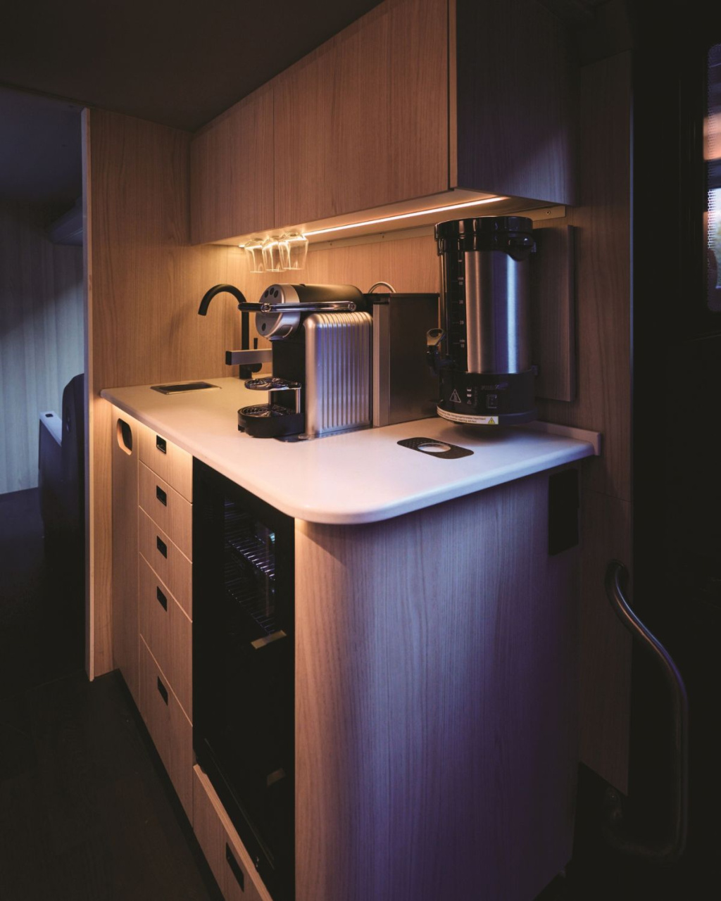 Compact luxury cabin kitchenette with wood cabinetry, a white countertop, espresso machine, and beverage dispenser under warm lighting.