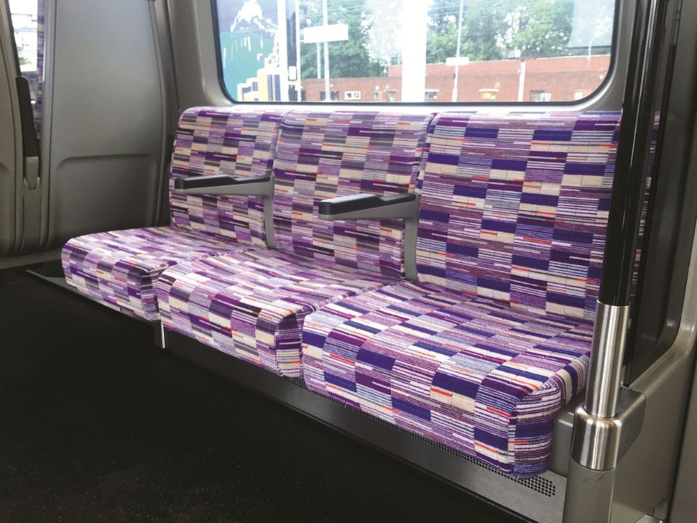 A clean train carriage with a long, empty bench seat upholstered in colorful purple, pink, and gray geometric stripes beneath a large window.