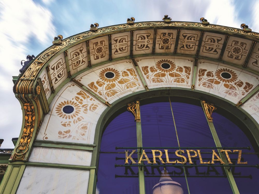 Ornate Karlsplatz entrance arch in Vienna, with cream-and-gold floral and geometric panels framing a deep blue glass sign against a cloudy sky.