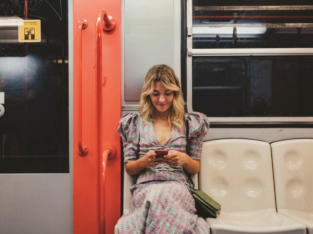 A woman in a pink-and-green plaid dress sits alone on a subway seat, smiling as she looks at her phone.