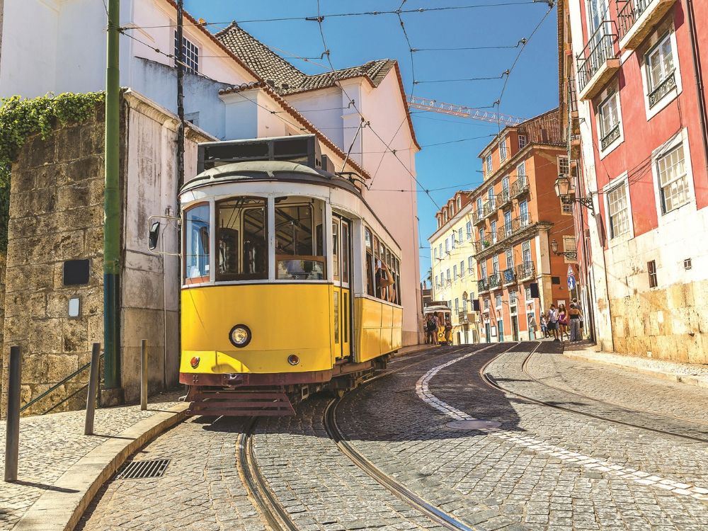 A bright yellow vintage tram curves along cobblestone tracks between colorful Lisbon buildings, beneath overhead wires on a sunny, lively street.