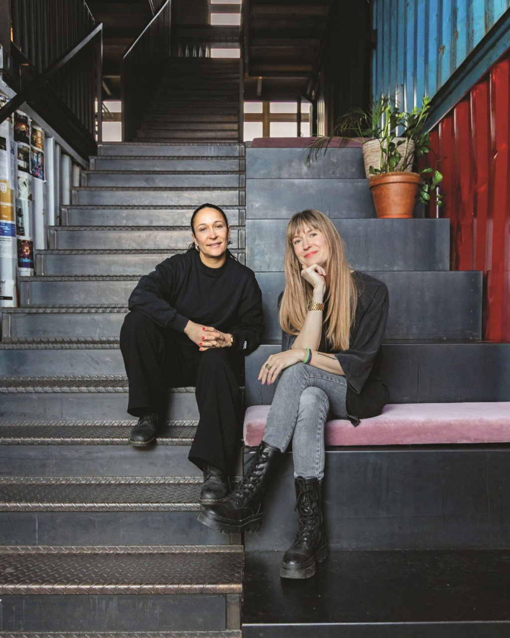 Two women sit on metal stairs in an industrial interior, one in black and one in gray with boots, beside a potted plant; their relaxed expressions feel warm and informal.