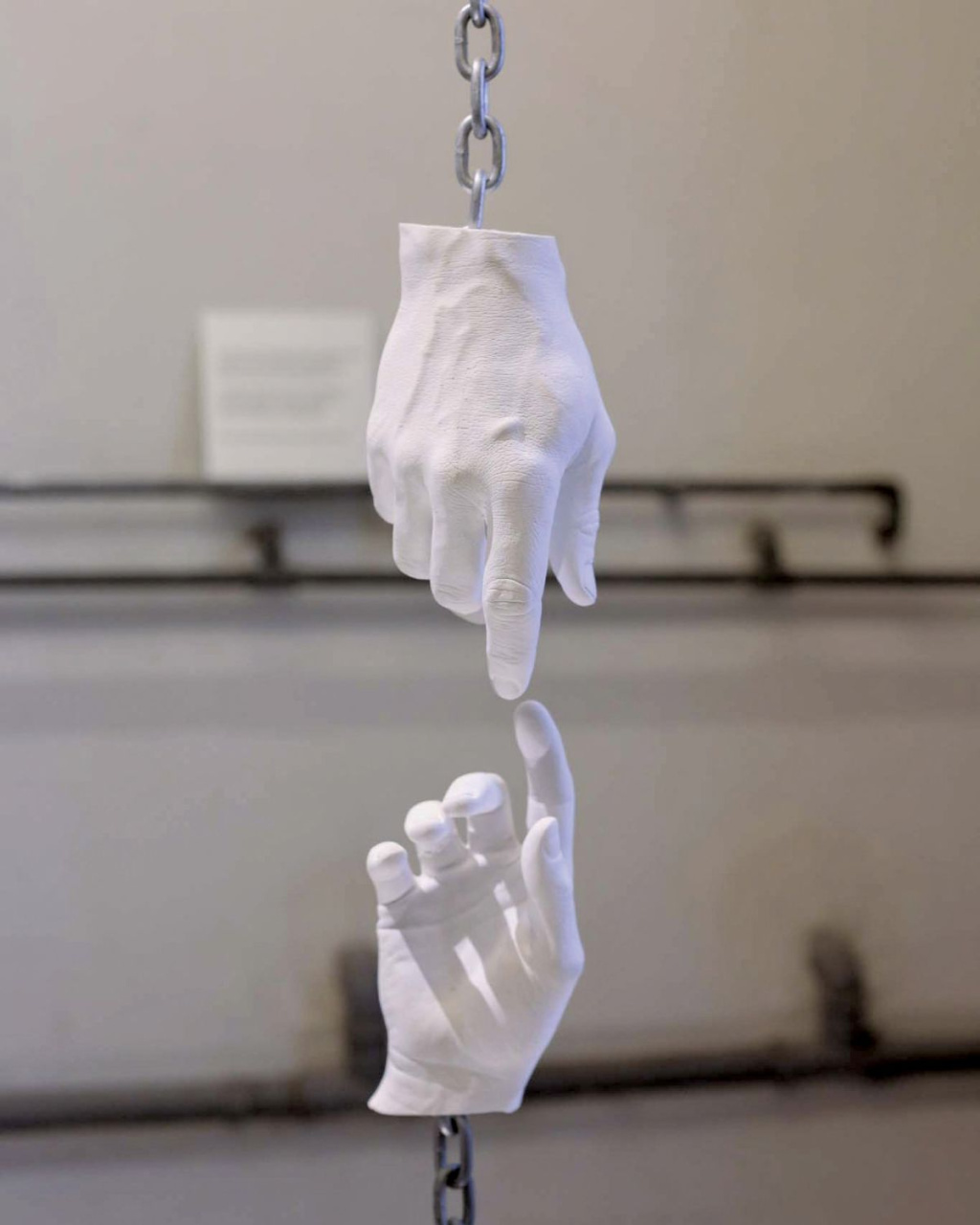 White sculpted hands hang from chains, one pointing down toward another reaching up, nearly touching against a muted industrial wall; the scene feels surreal and contemplative.