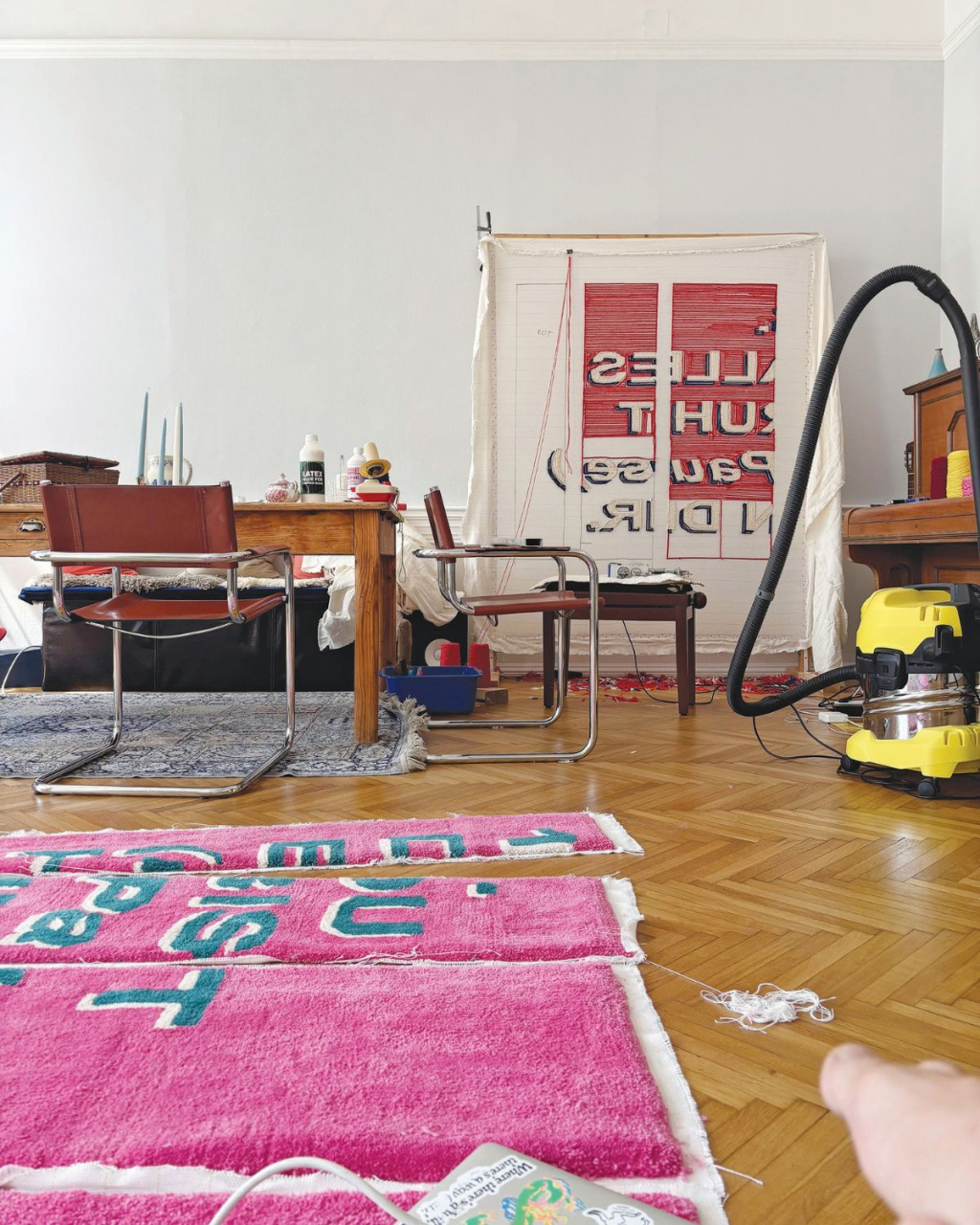 A cluttered indoor workspace with a worktable, chairs, vacuum, and tools around a large sign or canvas; bright pink rugs soften the busy, lived-in room.