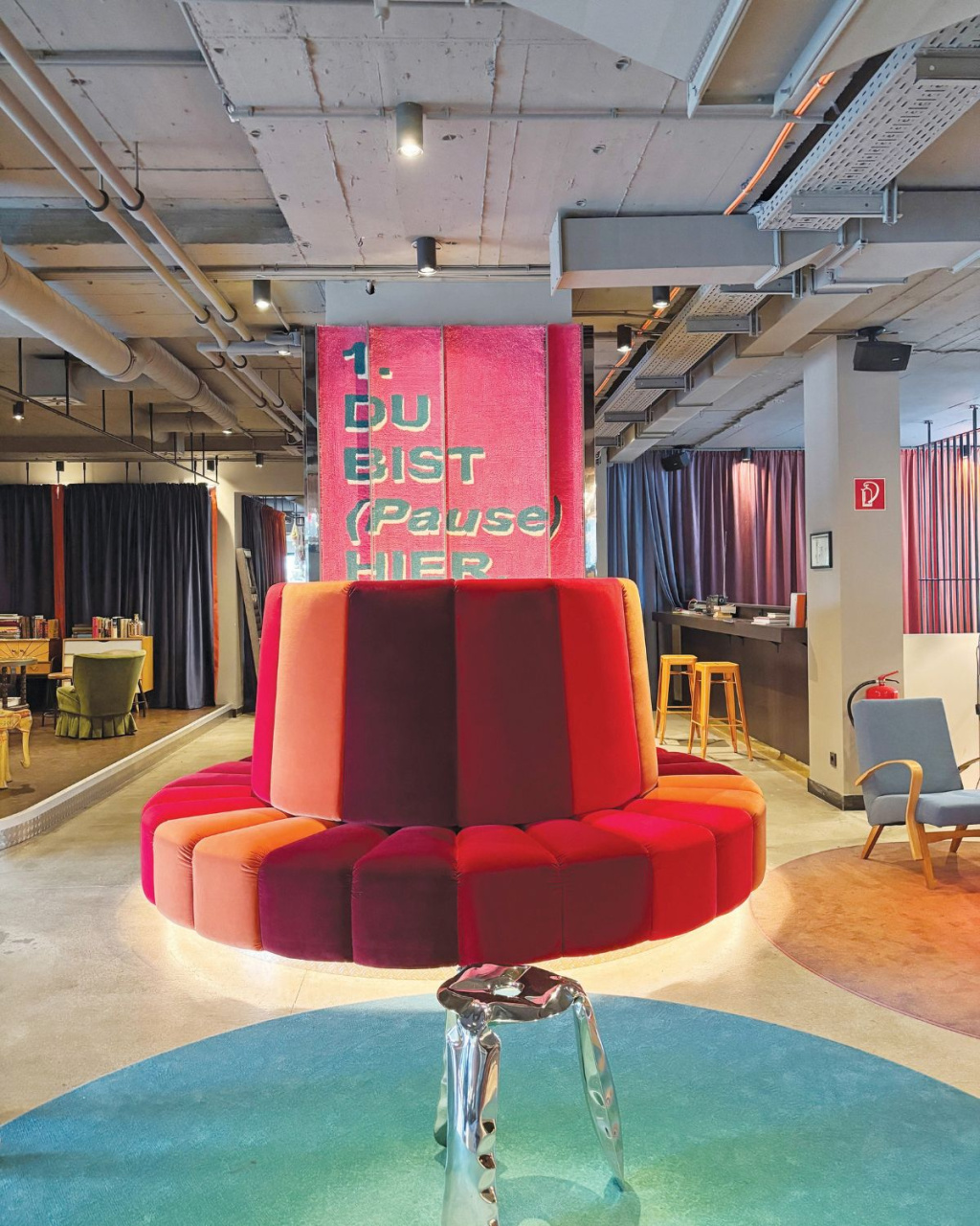 A vivid red-and-pink curved booth fills a modern lobby, beneath a playful German sign reading “Du bist (Pause) hier”; the space feels quirky and welcoming.