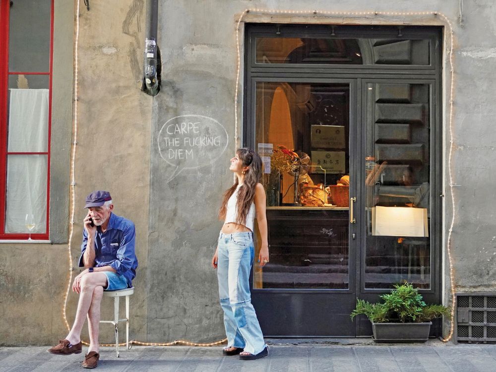 A woman stands beside a seated older man outside a warmly lit bakery, against a weathered wall with graffiti.